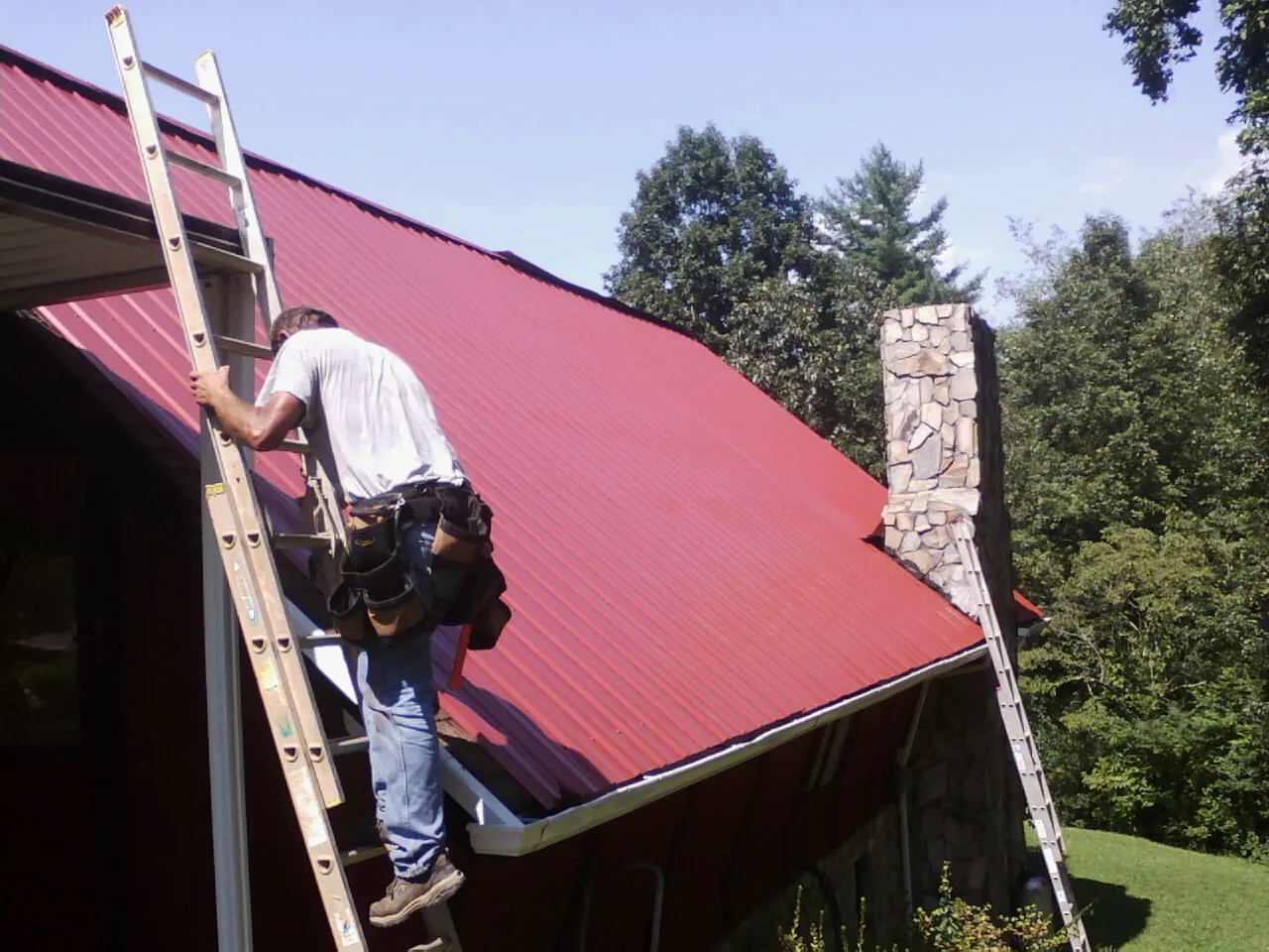 Roofer on a ladder beside a red metal roof and stone chimney — Soffit & Fascia Repair by Roofing Brothers in Clarendon Hills